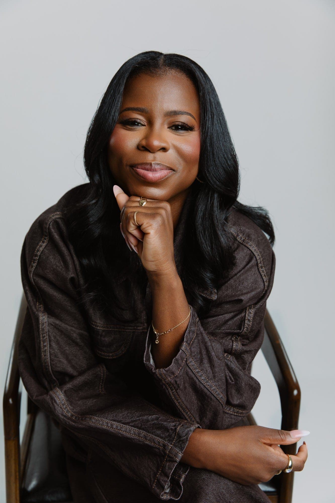 Manouchka Charles seated in a wooden chair, her chin resting on her hand, smiling gently, in a brown denim jacket against a pale grey studio backdrop.