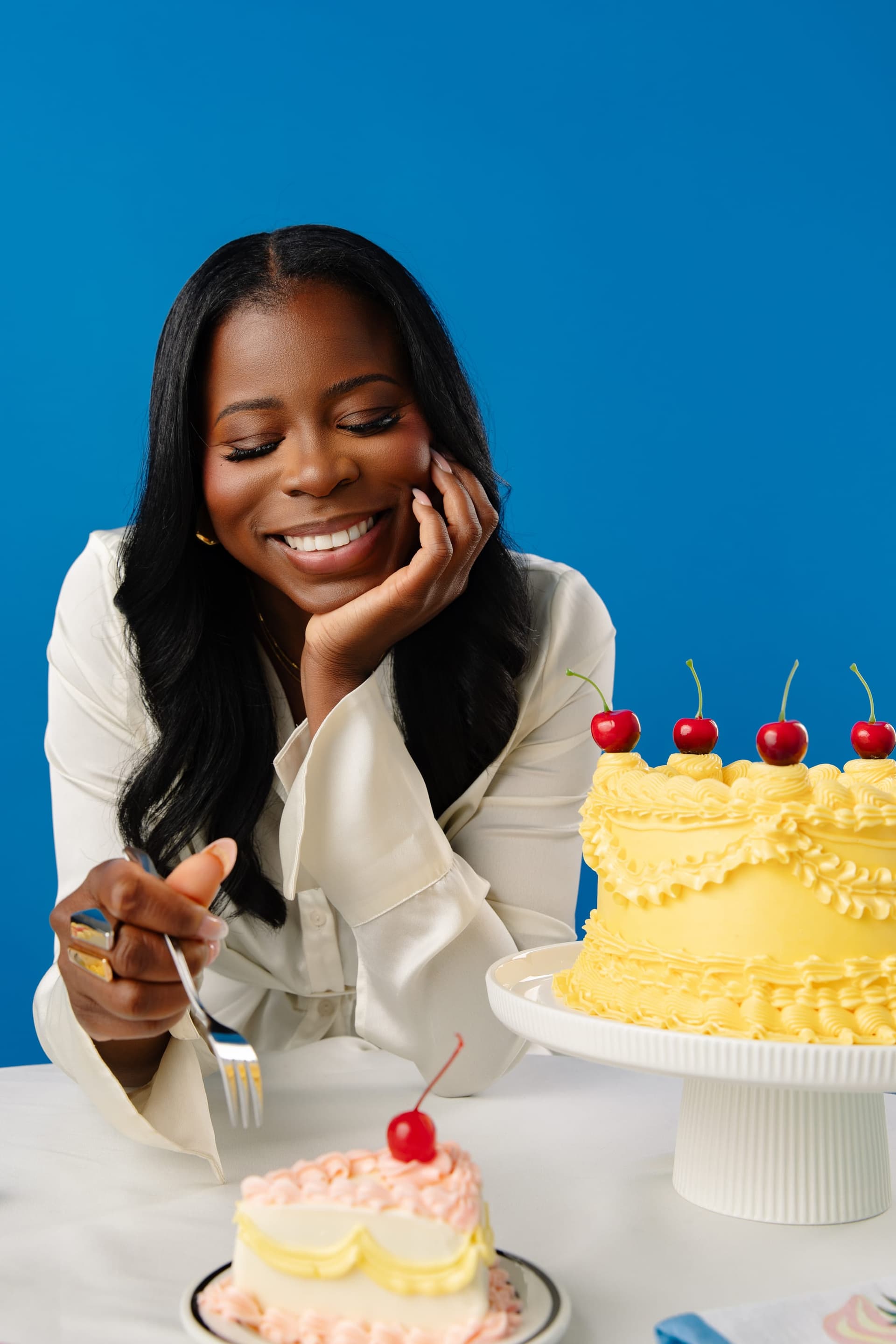 Manouchka resting her chin on her hand, smiling down at a yellow buttercream cake topped with cherries.