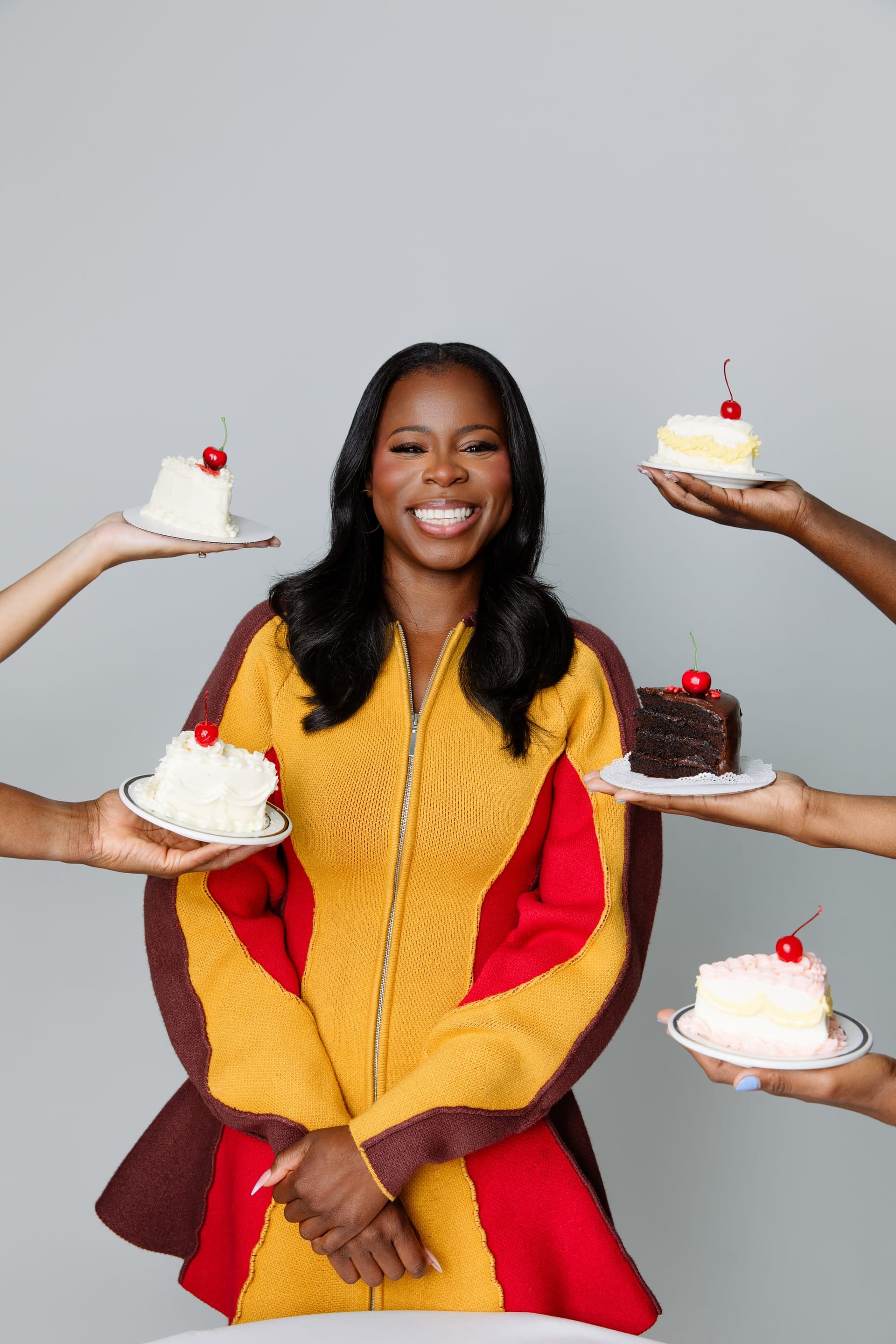 Manouchka Charles smiling to camera in a yellow and red coat, surrounded by five outstretched hands each offering her a small cherry-topped cake.