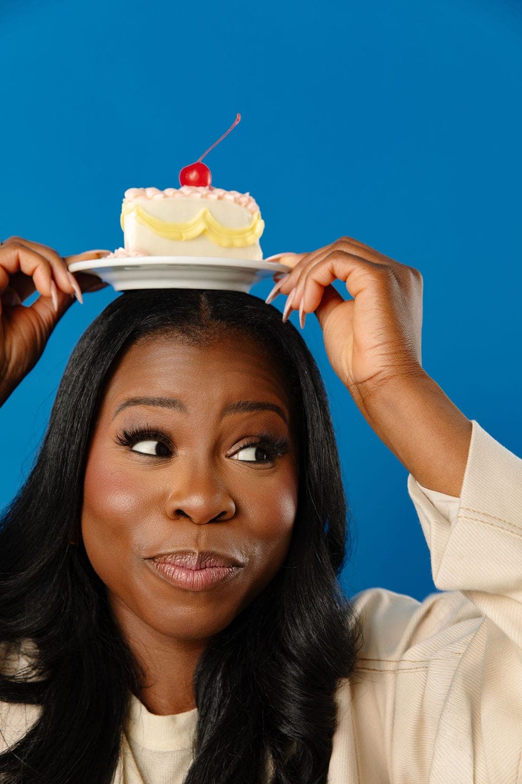 Manouchka Charles steadying a white plate on top of her head with both hands, a small cake with pink piping and a maraschino cherry balanced on it, glancing sideways with her lips pressed together in a wry look, against a cobalt blue backdrop.